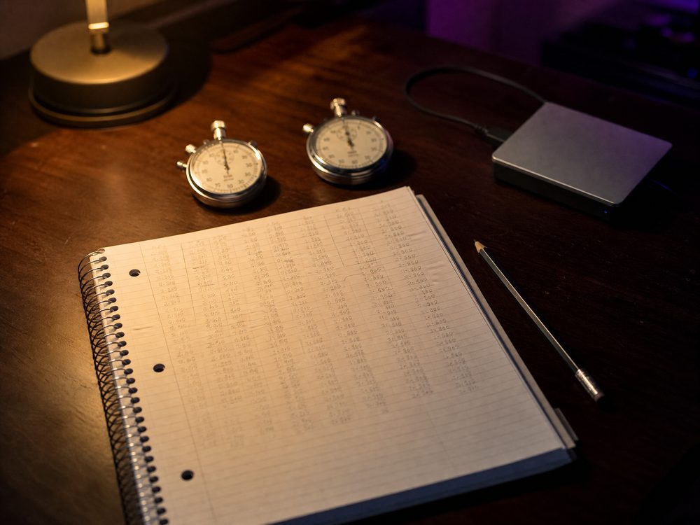 Two mechanical stopwatches beside a spiral notebook filled with columns of handwritten times on a dark walnut desk under a warm lamp